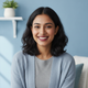 Smiling young South Asian woman in a casual top, with a warm and trustworthy expression against a soft sky blue background.